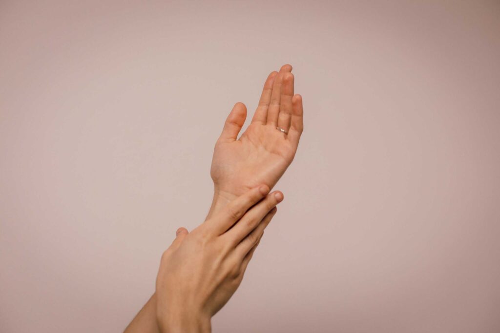 A hand is gently holding and supporting the wrist of another hand against a plain background.