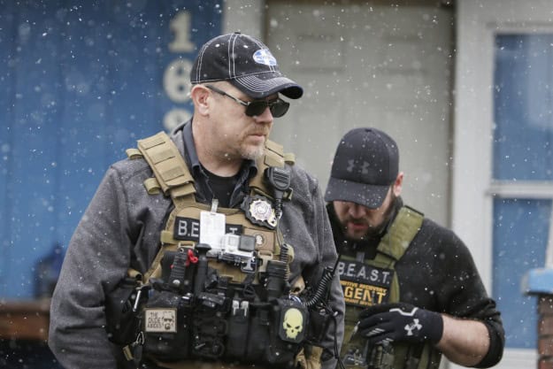 Two law enforcement officers wearing tactical gear stand outside in the snow.