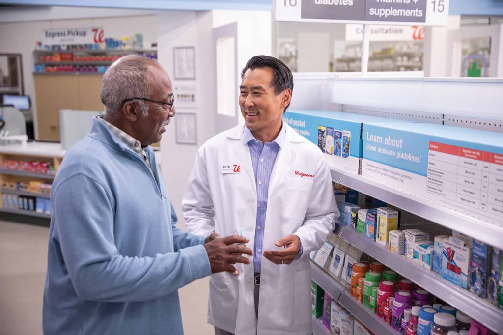 A pharmacist talks with a customer in a pharmacy aisle next to shelves stocked with medication.