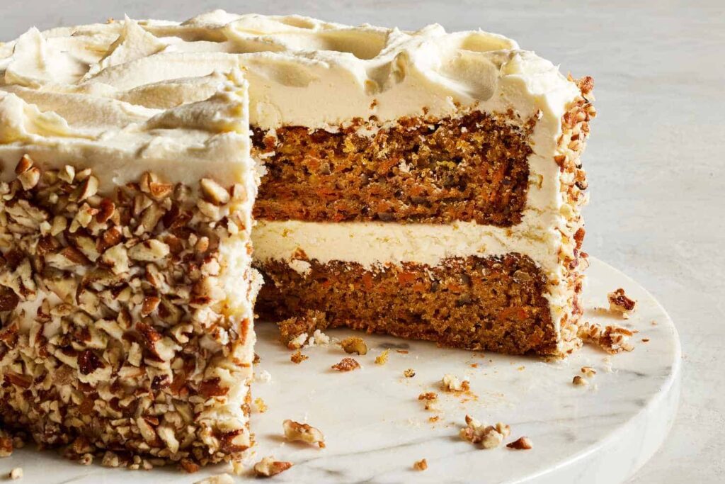 Carrot cake with cream cheese frosting and chopped nuts on the sides, with a slice missing, on a marble surface.