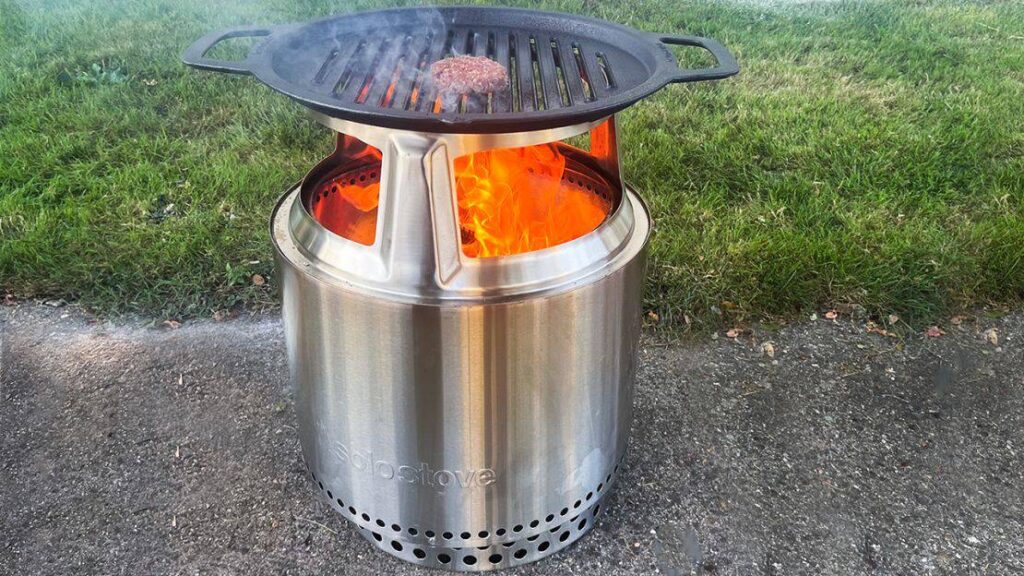 A stainless steel portable fire pit with a grill attachment on top, cooking a single burger patty over bright, open flames. Set on a concrete surface near grass.