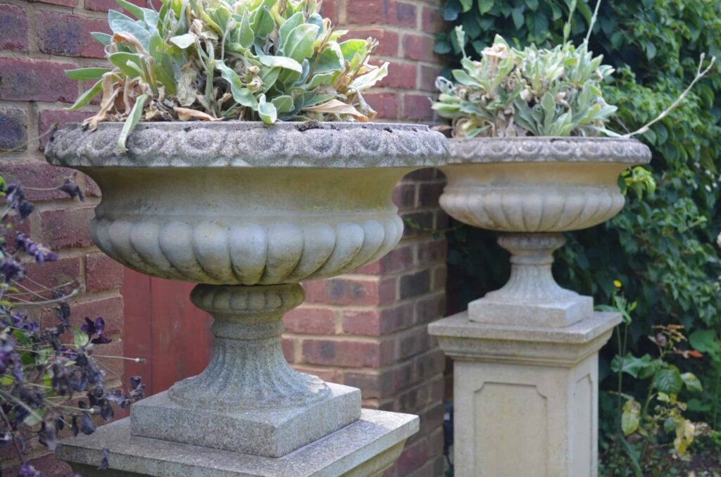 Two stone planters with ornamental designs hold leafy plants. They are placed on square pedestals against a brick wall.