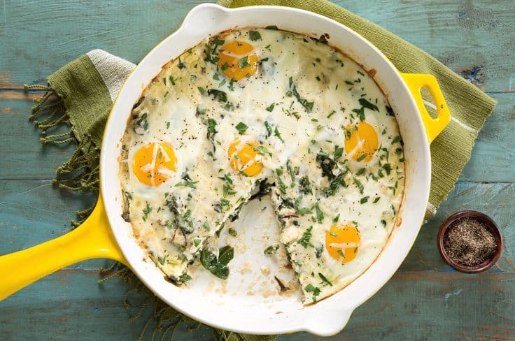 A skillet with baked eggs, spinach, and herbs on a wooden table. A slice is missing from the dish.