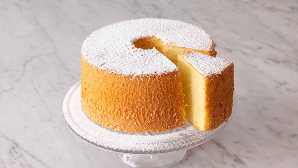 A round sponge cake with powdered sugar on top sits on a glass cake stand, missing one slice.