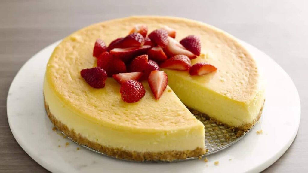 A whole cheesecake with a slice removed, topped with fresh halved strawberries, on a round white plate.