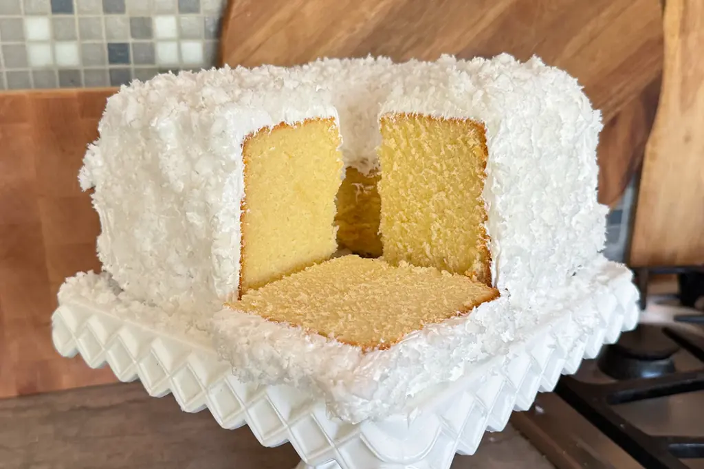 Square coconut cake with a slice removed, revealing yellow interior, on a white serving dish.