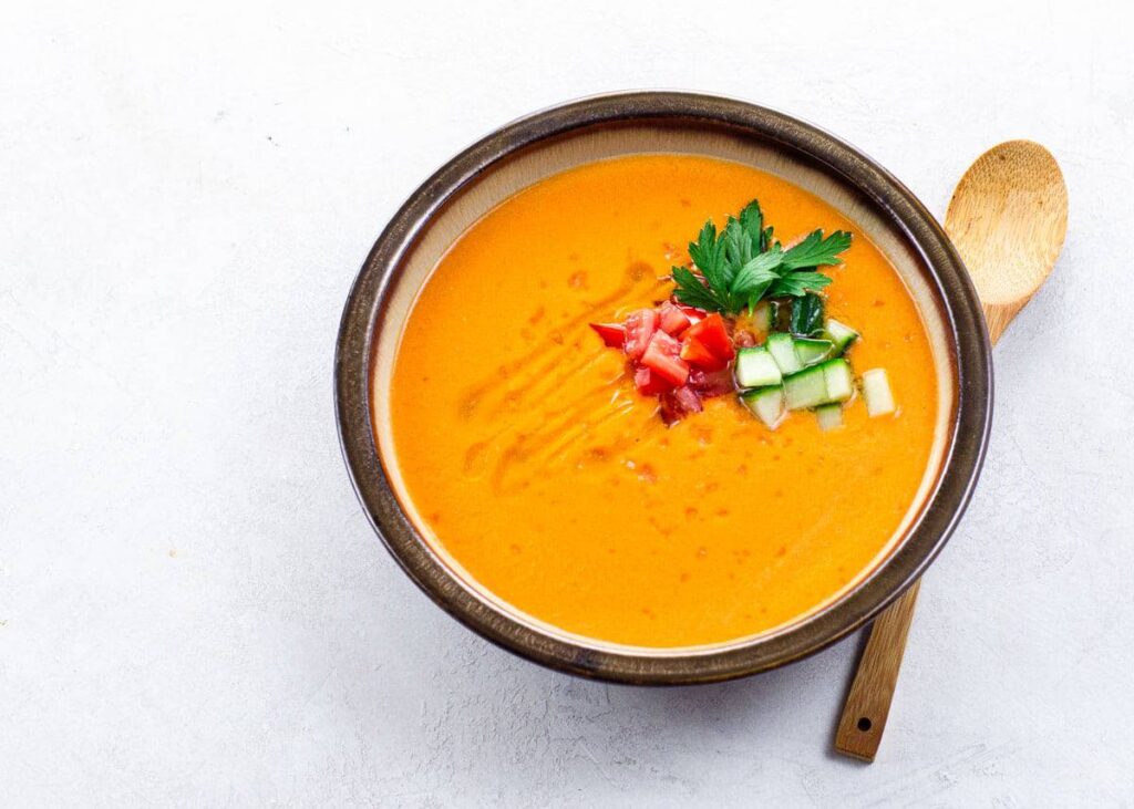 A bowl of orange soup garnished with diced tomatoes, cucumber, and parsley, with a wooden spoon on the side.