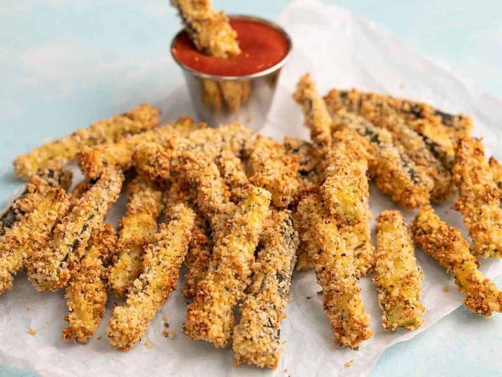 Crispy baked zucchini fries on parchment paper with a small cup of marinara sauce.