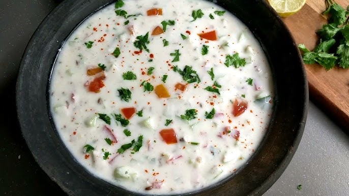 A black bowl filled with raita, featuring a creamy yogurt base mixed with diced vegetables and herbs. Lime and cilantro garnish are seen on the side.