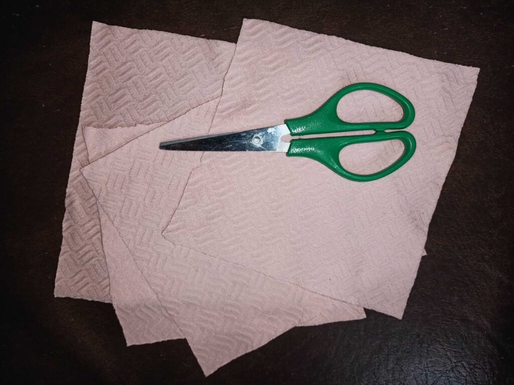 A pair of scissors with green handles rests on four textured beige cloths placed on a dark surface.