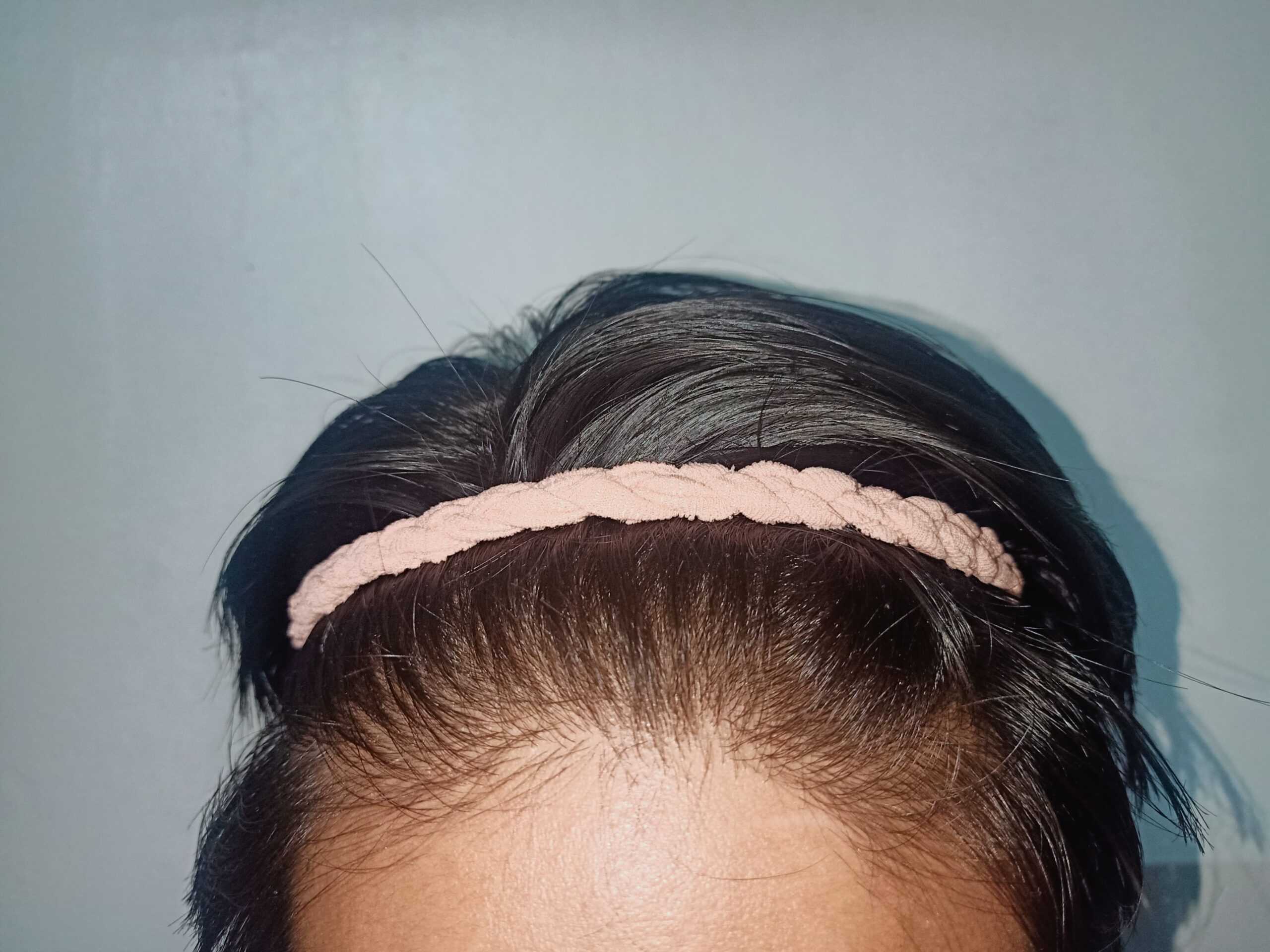 A person wearing a pink braided headband on their short, dark hair against a light blue background.