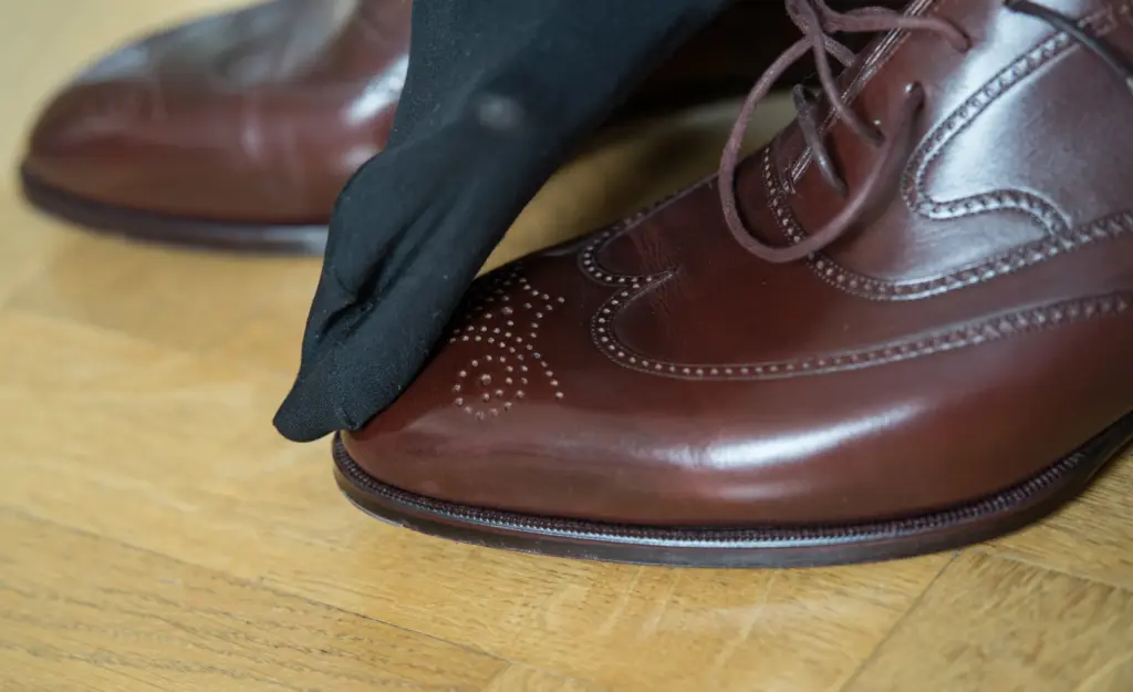 A hand in a black glove is polishing a brown leather dress shoe on a wooden floor.