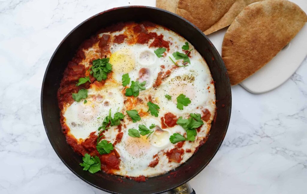 A skillet with shakshuka, topped with fresh parsley, next to two pieces of pita bread on a white surface.