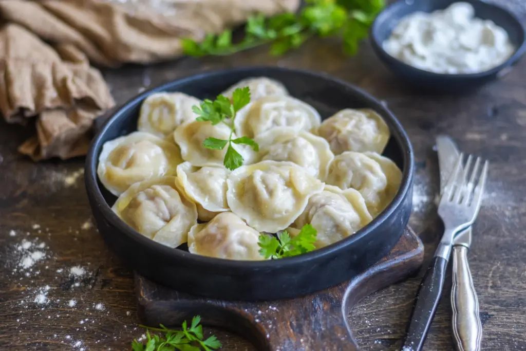 A black bowl filled with dumplings garnished with fresh parsley, placed on a wooden board. A fork and knife are beside the bowl, and a bowl of sauce is in the background.