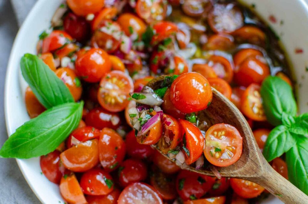 A wooden spoon holds cherry tomatoes, onions, and herbs over a bowl of the same mixture, garnished with fresh basil leaves.