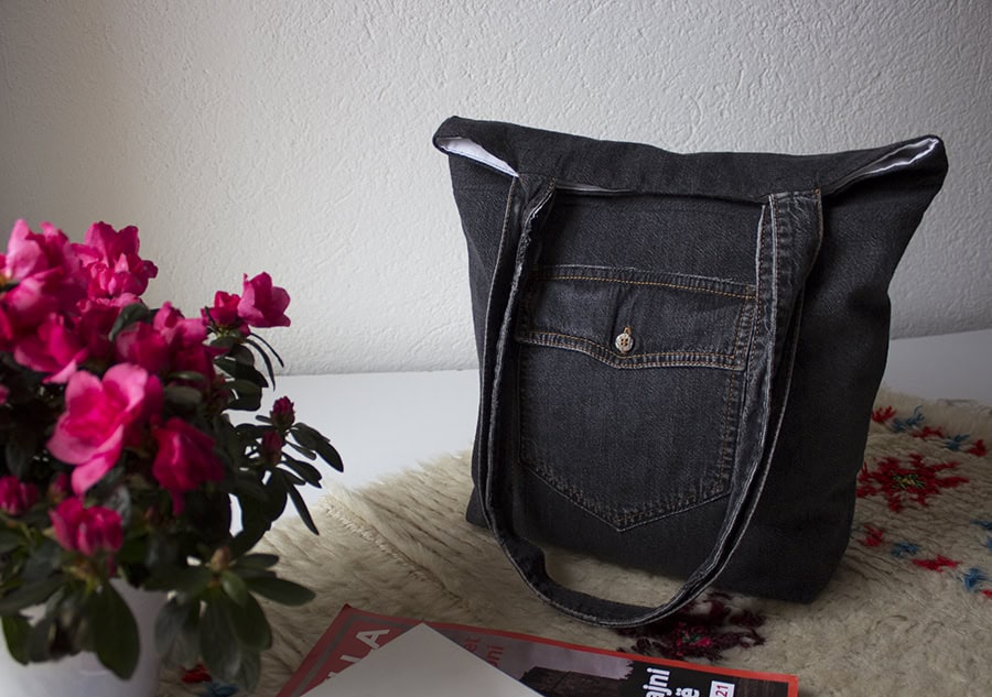 A denim tote bag with a front pocket is placed on a table next to a pot of pink flowers and a partially visible magazine.