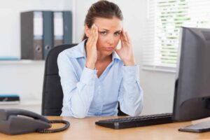 A woman in a blue shirt sits at a desk with a phone and keyboard, holding her head with both hands and looking at a computer screen, appearing stressed.