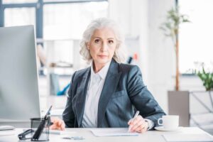 Elderly woman in a suit sitting at a desk with papers and a pen, looking at the camera. A computer monitor and a cup are on the desk. Office background with bright lighting.