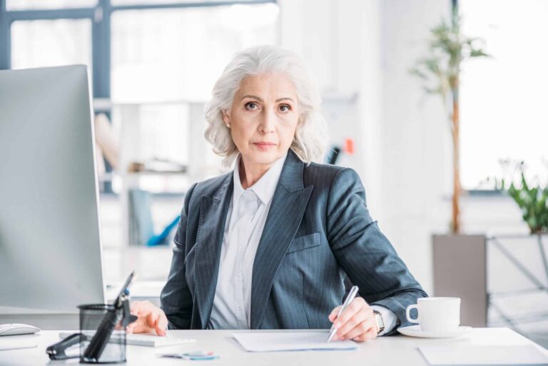 Elderly woman in a suit sitting at a desk with papers and a pen, looking at the camera. A computer monitor and a cup are on the desk. Office background with bright lighting.