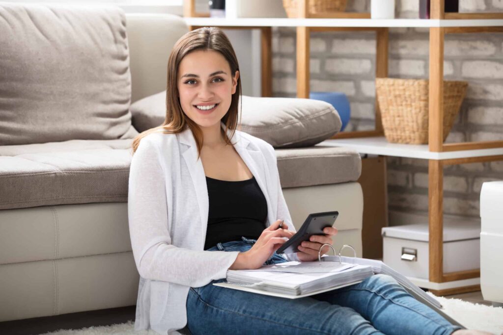 A person sits on the floor, smiling, holding a smartphone. Nearby are open folders and a couch.