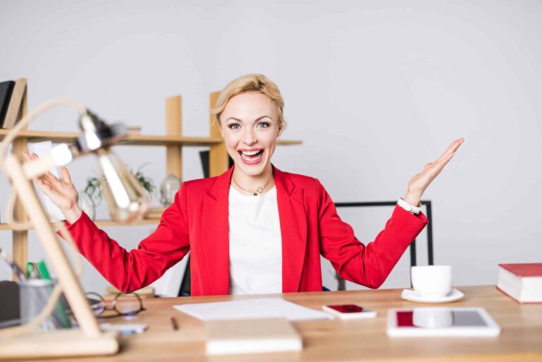 Person in a red blazer sits at a desk with open arms and a cheerful expression, surrounded by office items like a lamp, books, a smartphone, and a cup.