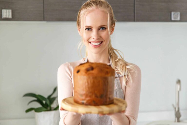 A person in an apron holds a freshly baked panettone on a wooden plate, smiling. A plant is in the background on a kitchen counter.