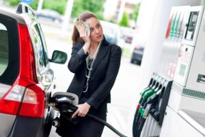A woman in a business suit stands at a gas pump, holding a nozzle with one hand and wiping her forehead with cash in the other. A car is parked next to her.
