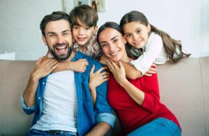 A family of four, including a man, woman, and two children, sit closely together on a couch, smiling at the camera.