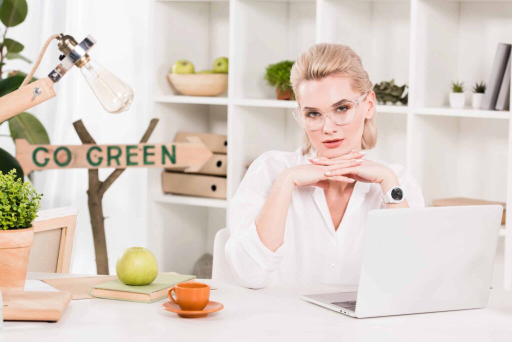 A person in glasses sits at a desk with a laptop, books, and a green apple. A "GO GREEN" sign and potted plants are in the background.