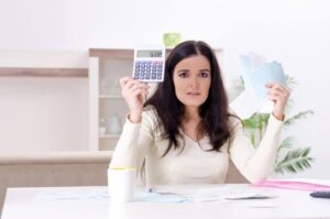 A woman sits at a table with a concerned expression, holding a calculator and several papers. A cup is on the table.