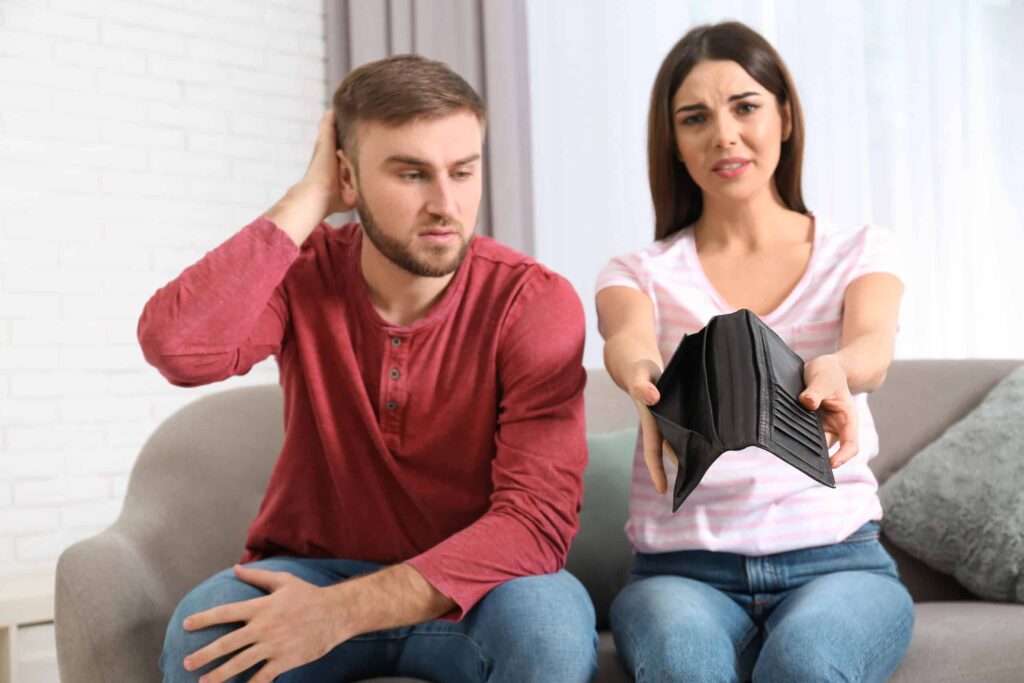 A couple sits on a couch; the woman holds an open, empty wallet while the man scratches his head.