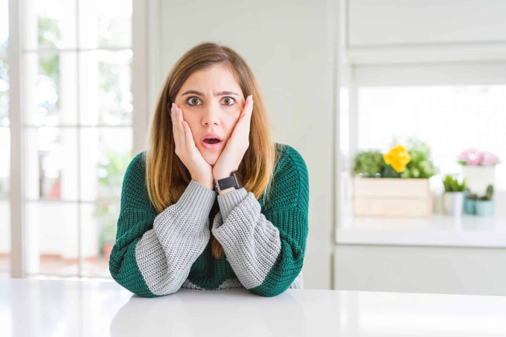 A woman with long hair sits at a table, resting her hands on her cheeks, looking surprised. She is wearing a green and gray sweater. The background shows a bright kitchen with plants.