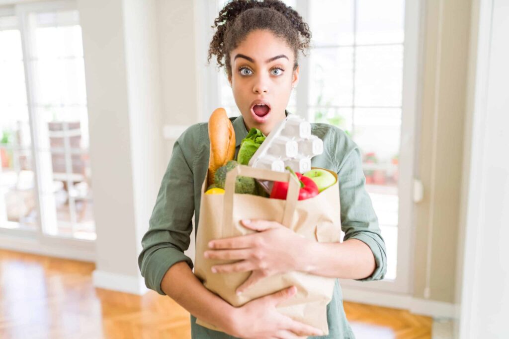 A person looks surprised while holding a paper bag filled with groceries, including bread, eggs, and vegetables, inside a bright room.