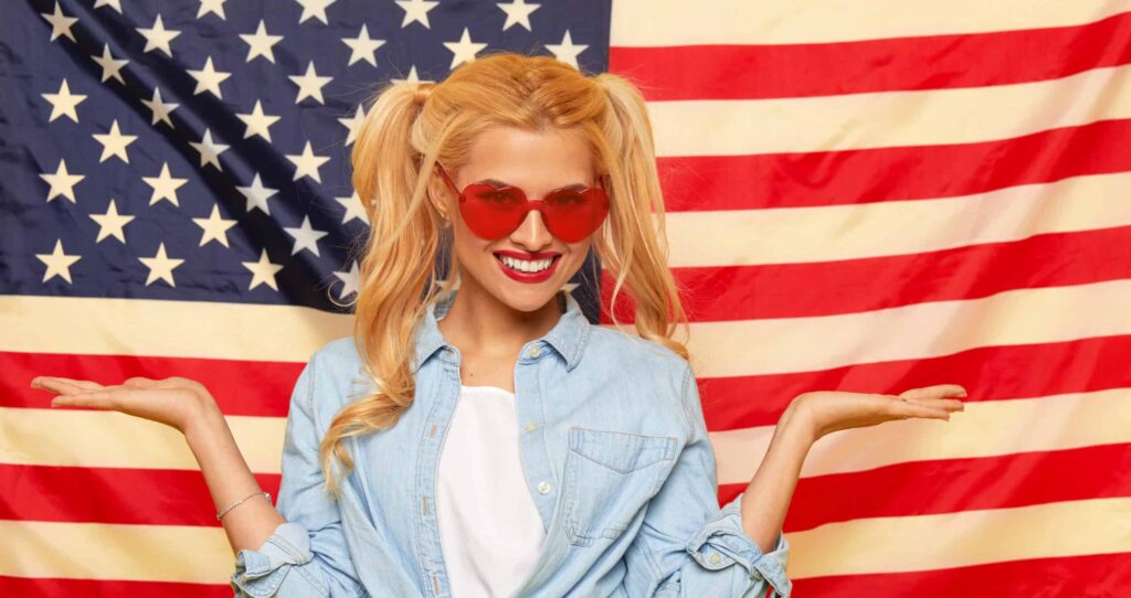 A woman with blonde hair and red heart-shaped sunglasses smiles, standing in front of a U.S. flag. She wears a denim shirt and white t-shirt, with her hands raised in a shrugging gesture.
