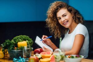 Person smiling while holding a pen and notebook, surrounded by fruits and vegetables on a kitchen counter.