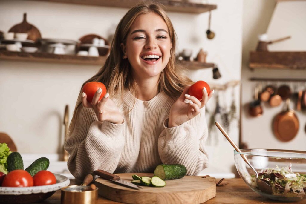 A woman holds two tomatoes while smiling at a kitchen counter with sliced cucumbers and a salad bowl.