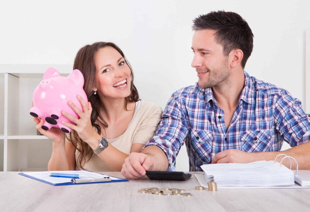 A smiling woman holds a piggy bank, sitting next to a man with a calculator. They appear to be budgeting with papers and coins on the table.