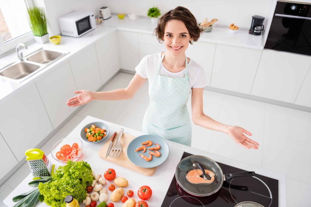 A woman in a light blue apron stands smiling in a modern kitchen. Fresh vegetables and a plate of shrimp are on the counter, while a pan of shrimp cooks on the stove.