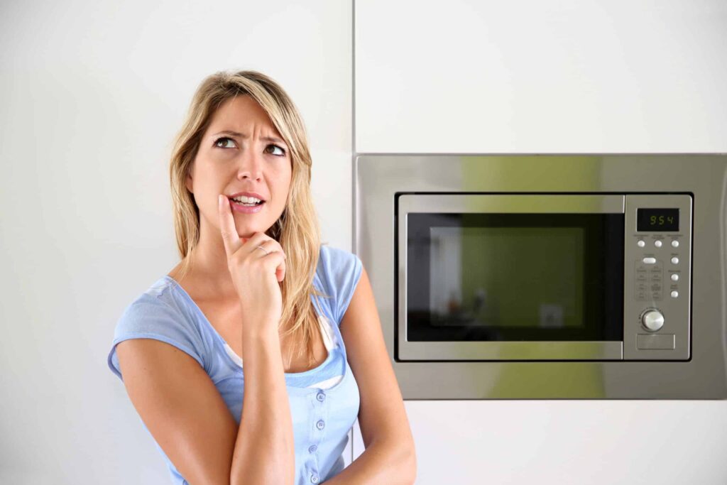 Person standing thoughtfully near a microwave oven, touching their chin.