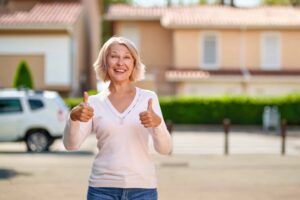 Elderly woman smiling, giving two thumbs up, standing outside with houses and a white car in the background.