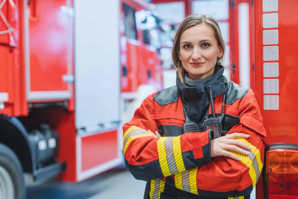 A firefighter in red and yellow gear stands with arms crossed in front of emergency vehicles.