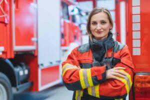 A firefighter in red and yellow gear stands with arms crossed in front of emergency vehicles.