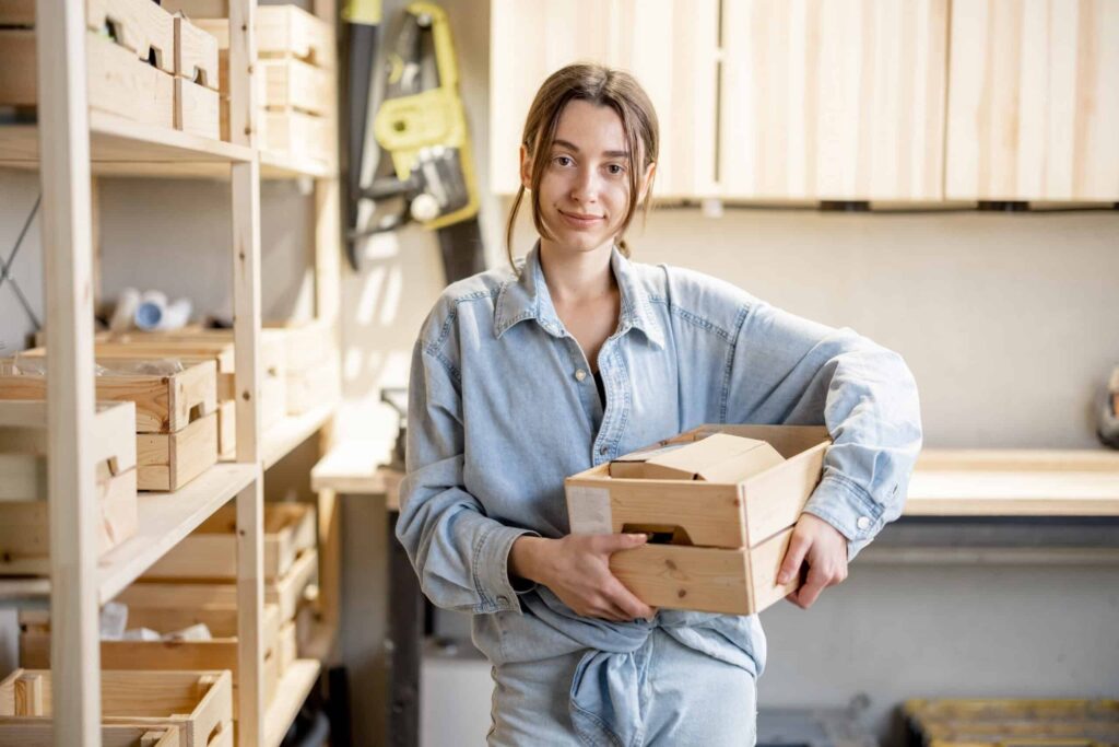 Person wearing denim shirt holds a wooden crate in a workshop with shelves containing wooden items in the background.