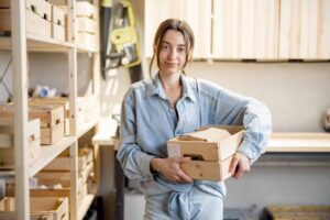 Person wearing denim shirt holds a wooden crate in a workshop with shelves containing wooden items in the background.
