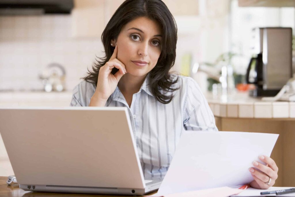 A woman sitting at a desk with a laptop, holding and looking at a piece of paper.