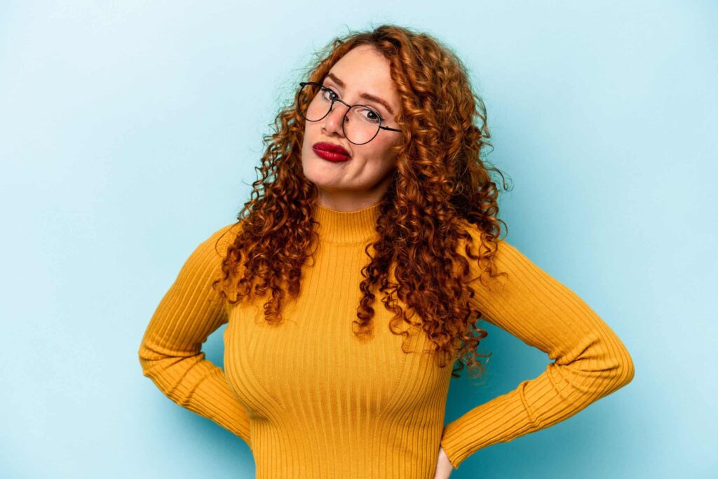 A person with curly red hair and glasses wears a mustard yellow sweater and stands against a light blue background with hands on hips.