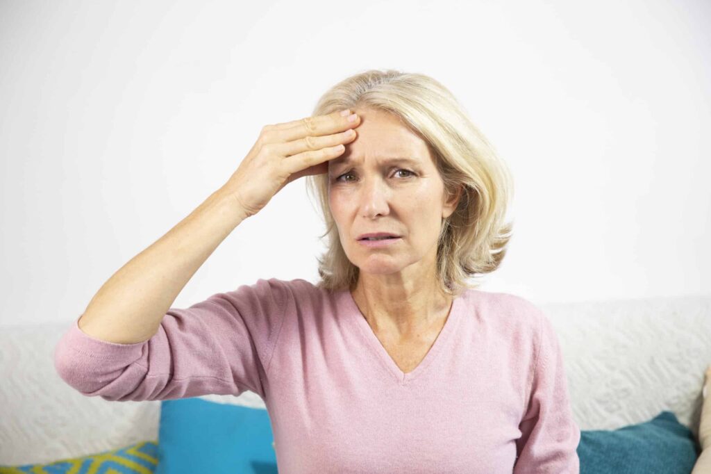 A woman with shoulder-length blonde hair wearing a pink sweater rests her hand on her forehead, looking concerned while sitting on a couch.