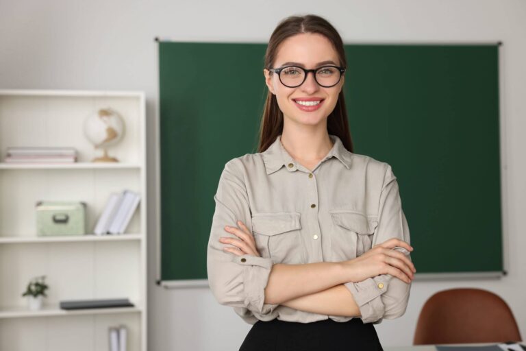 A person with glasses stands smiling with arms crossed in a classroom. A chalkboard is in the background, and shelves with books and a globe are on the left.