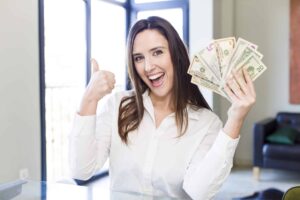 Person in a white shirt smiling, holding a fan of cash in one hand, and giving a thumbs-up with the other, in a bright room.