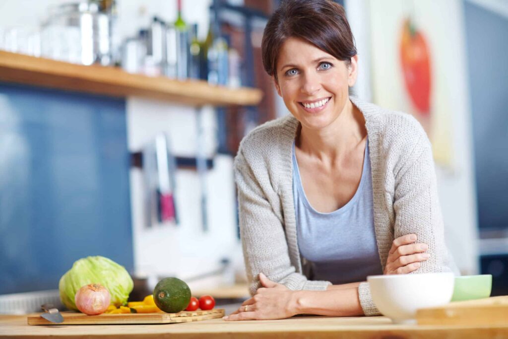 Woman smiling in a kitchen with vegetables on the counter, including an avocado, tomato, and lettuce.